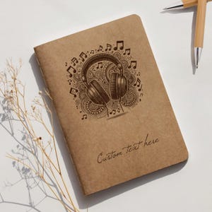 Op de afbeelding: Een bruin notitieboek met een muzikaal thema. De omslag toont een gedetailleerde illustratie van een koptelefoon omringd door muzieknoten en decoratieve krullen. De tekst "Custom Text here" staat onderaan.