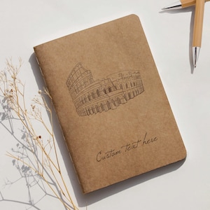 Op de afbeelding: Een bruin notitieboek met een schets van het Colosseum en de tekst "Custom Text here" in cursief. Twee pennen zijn zichtbaar op de achtergrond. Het notitieboek ligt op een wit oppervlak.