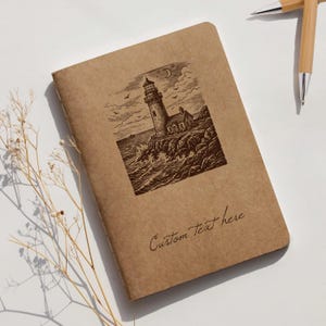 Op de afbeelding: Een bruin notitieboek met een gedetailleerde illustratie van een vuurtoren op een rotsachtige kust, met golven die breken. De tekst "Custom Text here" is cursief geschreven. Twee bamboe pennen staan op de achtergrond.