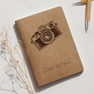 Puede incluir: Un cuaderno marrón con una ilustración de cámara vintage. El cuaderno tiene la frase "Custom Text here" impresa en la portada. Dos bolígrafos de bambú en el fondo.
