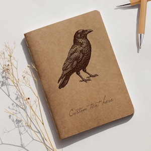 Könnte beinhalten: Ein braunes Notizbuch mit einer detaillierten Illustration einer Krähe. Die Krähe ist im Vintage-Stil dargestellt. Die Worte "Custom Text here" sind kursiv am unteren Rand des Notizbuchs geschrieben.
