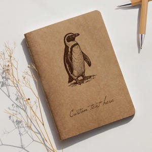 May include: A brown notebook featuring a detailed penguin illustration. The notebook has the words "Custom Text here" written in cursive. Two bamboo pens are in the background.