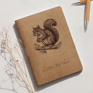 Puede incluir: Un cuaderno marrón con una ilustración detallada de una ardilla sosteniendo una nuez, posada en una rama. El texto "Custom Text here" está escrito en cursiva. Dos bolígrafos de bambú están en el fondo.
