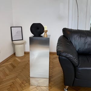 May include: A tall, rectangular, metallic silver pedestal table with a gradient finish. A black, circular, textured decorative object and a small wooden candle holder with a white candle sit on top. A black leather sofa and a silver floor lamp are in the background.