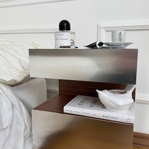 Può includere: Un comodino moderno con finitura metallica, con una bottiglia di profumo, una tazzina e un piattino, e un tubetto di crema. Sono presenti anche un uccello in ceramica bianca e un libro con la scritta "GOOD VIBES, GOOD LIFE".