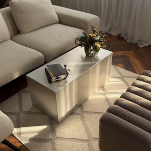 May include: A contemporary living room with a brushed metal coffee table, a black notebook, and a cream-colored tube. A beige sofa, patterned rug, and brown ottoman are also visible. A vase of flowers sits on the table.