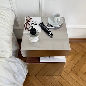 May include: A modern, two-tiered side table with a brushed metal top and a warm brown base. The top surface holds a white teacup and saucer, a perfume bottle, a tube of cream, and eyeglasses. A book is visible on the lower shelf.