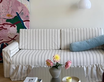 May include: A sofa with a white and black vertical striped fabric cover and a ruffled skirt, featuring a light blue bolster pillow. A square metallic coffee table holds a gray vase with pink flowers, a stack of books, and a yellow cup on a tray.