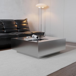 May include: A modern, square, silver coffee table with a low profile. The table is in a living room setting with a black leather sofa, a floor lamp, and a rolled-up magazine. The table is approximately 36 inches square.