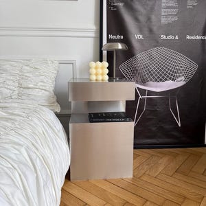 Può includere: Un comodino in metallo spazzolato con ripiano aperto, dotato di una candela a bolle color crema e una piccola lampada argentata sopra. Un libro nero poggia sul ripiano. Accanto c'è un grande poster nero con testo bianco e il grafico di una sedia.