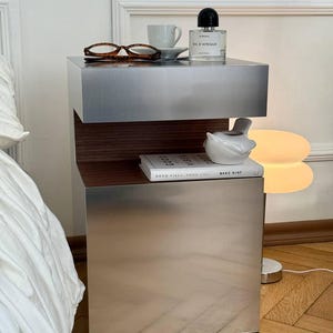 Puede incluir: Una mesita de noche moderna y metálica con un diseño minimalista. La mesa presenta dos secciones rectangulares apiladas con un interior de madera. Una taza, gafas, perfume y un libro se exhiben en la parte superior. También se ve una figura de pájaro de cerámica y una lámpara.