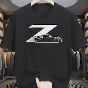 Peut inclure: T-shirt noir avec un motif blanc représentant une voiture stylisée et la lettre "Z". Le design de la voiture comprend des lignes de vitesse, suggérant le mouvement. Le t-shirt est suspendu à un cintre en bois.