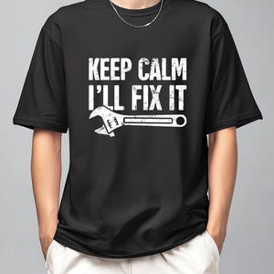 May include: Black t-shirt with the white text "KEEP CALM I'LL FIX IT" and a white wrench graphic. The shirt is a crew neck style and appears to be made of a soft material. The design is centered on the front of the shirt.