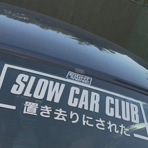 Op de afbeelding: Witte sticker op een autoruit met de tekst "SLOW CAR CLUB" in grote, vette letters. Japanse karakters zijn ook zichtbaar. De sticker heeft ook het Eight Nine logo.