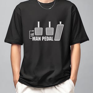 Może przedstawiać: Czarny t-shirt z białym nadrukiem trzech pedałów samochodowych i napisem "THE MAN PEDAL". Projekt zawiera strzałkę. Odzież codzienna.