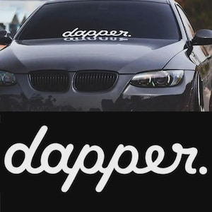 May include: A white vinyl car sticker with the word "dapper" in a cursive font is displayed on the windscreen of a dark grey car. The sticker is centred and has a clean, modern aesthetic. The car's grill and headlights are visible.