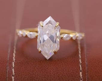 May include: A gold ring with a large, clear, hexagonal-cut gemstone as the centerpiece. The band features smaller, clear gemstones. The ring is set against a brown leather background.
