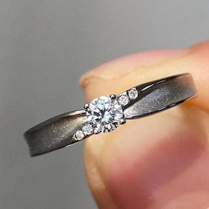 Puede incluir: Un anillo plateado con una piedra preciosa central redonda y transparente y piedras de acento más pequeñas. La banda del anillo tiene un acabado texturizado. El anillo se sostiene entre los dedos sobre un fondo borroso.