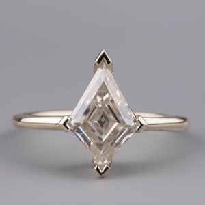 May include: A silver ring with a kite-shaped diamond. The clear gemstone is the centrepiece, with a unique cut and sparkle. The ring has a thin band and decorative accents on the setting. The ring is set against a grey background.