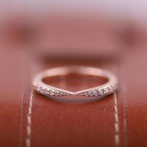 May include: A delicate rose gold band with a row of small diamonds set in a V-shape. The ring is on a brown leather surface.