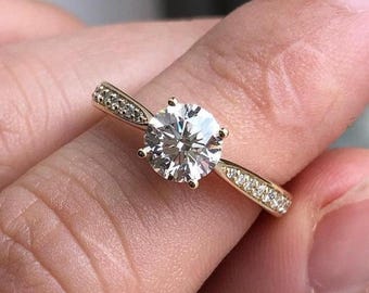 May include: A gold engagement ring with a large, round, clear diamond center stone. The band is also gold and features smaller accent stones. The ring is held up by a finger.