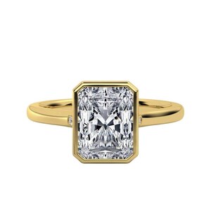 May include: A radiant-cut diamond ring with a gold band. The diamond is set in a bezel setting, and small accent diamonds are on each side of the main stone. The ring is set against a white background.