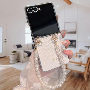 Könnte beinhalten: Eine weiße Handyhülle mit Perlenriemen. Die Hülle hat ein strukturiertes, schlangenlederartiges Muster und goldfarbene Hardware. Der Riemen besteht aus weißen Perlen. Das Telefon ist schwarz.