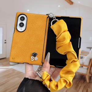 May include: A mustard yellow textured phone case with a matching fabric scrunchie wrist strap attached by a silver metal ring. The case features a small black flower emblem and a white number five charm with rhinestones on the back cover.