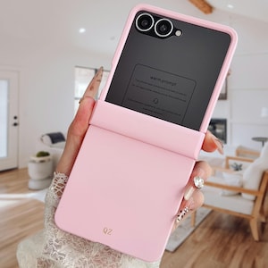 以下が含まれることがあります： 滑らかなマット仕上げのライトピンクの折りたたみ式スマホケースで、下部にゴールドでQZの文字がプリントされています。ケースには黒いスクリーンプロテクターエリアがあり、warm promptというテキストと説明が記載された長方形のラベルが付いています。