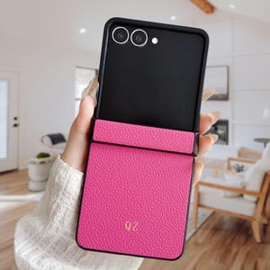 May include: A hot pink phone case with a black frame, open to reveal a black interior. The case has a textured finish and the initials "QZ" in gold. The phone's camera lenses are visible at the top.