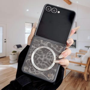以下が含まれることがあります： ヒョウ柄のデザインが施された、グレーと黒の携帯電話ケース。ケースにはシルバーのリングがあり、下部には「QZ」の文字がプリントされています。携帯電話ケースは開いており、携帯電話のカメラと画面が表示されています。