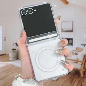 Peut inclure: Une coque de protection transparente pour smartphone pliable, dotée d'un anneau magnétique circulaire blanc sur le panneau arrière inférieur et d'un cache-charnière horizontal blanc. Le design est transparent avec les lettres QZ en doré.
