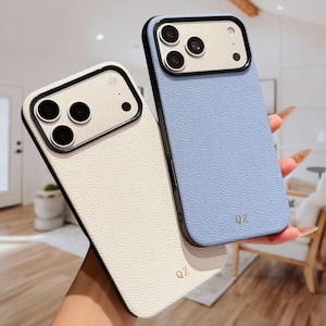 Puede incluir: Dos fundas para smartphone con acabado texturizado, una en blanco y otra en azul claro. Cada funda presenta un marco de cámara negro y las letras QZ en color dorado impresas cerca de la parte inferior. Las fundas tienen una textura granulada.