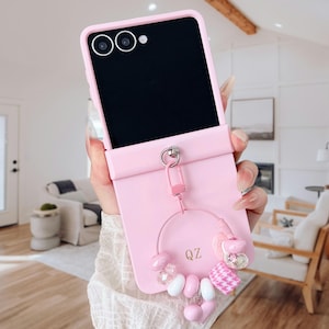 May include: A pink phone case with a decorative charm. The case is designed for a folding phone and features a loop for the charm. The charm includes beads, a heart, and a pink and white patterned fabric. The letters "QZ" are visible on the charm.