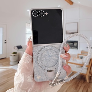 May include: A silver and grey phone case with a denim-like texture and a matching strap. The case is open, revealing a black screen and camera lenses. The strap is a braided silver cord.