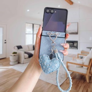Op de afbeelding: Een lichtblauwe denim telefoonhoes met een hartvormige bedel en polsband. De hoes heeft een zilveren hartvormige sluiting en een bijpassende denim hartbedel met pailletten accenten. De polsband is een lichtere tint blauw.