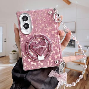 以下が含まれることがあります： ピンクのグリッター加工が施されたスマートフォンケースで、リボンのパターンと「DongSheng」の文字が入ったハート型のリングスタンドが付いています。小さなピンクの布製リボンが付いた取り外し可能なビーズストラップ、シルバーの金具、デュアルカメラ用の切り抜きが特徴です。