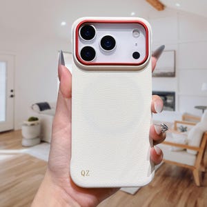 Puede incluir: Funda de teléfono blanca texturizada con un marco de cámara rojo. La funda tiene una textura similar al cuero y presenta tres lentes de cámara y un flash. Las letras "QZ" están impresas en dorado en la parte inferior de la funda.