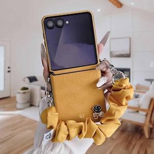 Peut inclure: Coque de téléphone jaune moutarde avec une dragonne scrunchie assortie. L'étui a une finition texturée et un cadre de caméra noir. Un petit charme de fleur noir et argent et le chiffre 5 sont attachés à l'étui. Les initiales "QZ" sont également visibles.