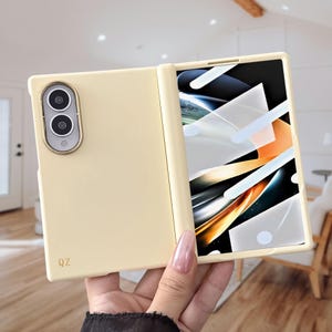 Könnte beinhalten: Eine cremefarbene, faltbare Smartphone-Hülle mit einem gold umrandeten Dual-Kamera-Ausschnitt auf der Rückseite und den in Gold gedruckten Buchstaben QZ. Die Hülle ist geöffnet und zeigt einen Bildschirm mit einer abstrakten Grafik aus weißen, orangefarbenen und schwarzen Formen.