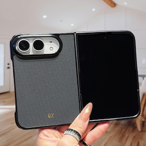 May include: A foldable smartphone case with a dark gray textured back panel featuring the gold letters QZ. The case has a black frame, a white camera module with two circular lenses, and an open right side revealing a black screen surface.