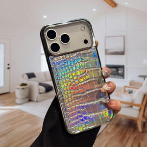 May include: A silver and iridescent phone case with a crocodile skin texture. The case has a silver frame and a clear camera lens protector. The iridescent finish reflects a rainbow of colours. The case is designed to fit a smartphone.
