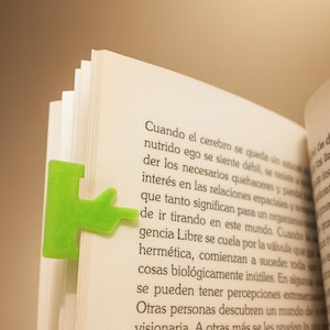 May include: A green, abstract-shaped bookmark is inserted into a book. The bookmark is made of a soft material and is a bright, eye-catching green. The book's pages are white, and text is visible.