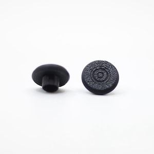 May include: Two black joystick thumb grips. One grip is shown from the side, revealing a cylindrical post. The other grip is shown from the top, with a textured surface featuring a circular pattern. The grips are set against a white background.
