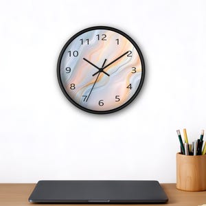 May include: A round wall clock with a black frame. The clock face features a swirling abstract design in shades of peach, blue, and white. Black hands indicate the time. The clock is hanging on a white wall above a wooden desk.