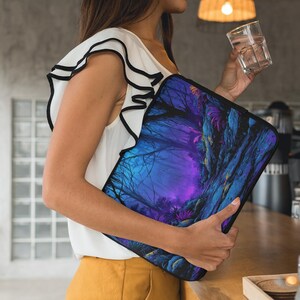 Puede incluir: Una funda negra para portátil con un diseño de bosque de fantasía en tonos azules, morados y verdes. La obra de arte representa árboles y plantas detallados, creando una escena mística. La funda es sostenida por una persona.