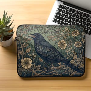 May include: A laptop sleeve featuring a detailed illustration of a crow perched on a branch, surrounded by floral and foliage elements. The design incorporates shades of blue, green, and cream, creating a nature-inspired aesthetic. The sleeve is placed on a wooden surface.