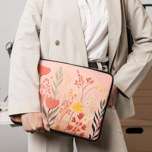 Puede incluir: Una funda para portátil con un diseño floral en tonos melocotón, rojo, amarillo y verde. La funda tiene un borde negro. La persona que sostiene la funda lleva una blazer beige.