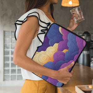 May include: A laptop sleeve featuring a colorful cloud design in shades of purple, blue, yellow, and orange. The sleeve has a black border and is held by a person wearing a white top and yellow pants.