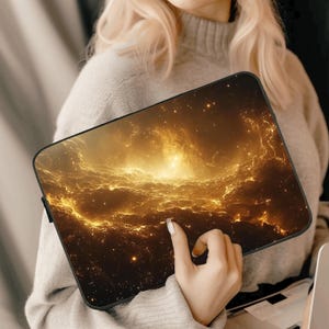 Op de afbeelding: Een zwarte laptophoes met een galaxy-thema. De hoes heeft een draaiend patroon van gouden en witte wolken tegen een donkere achtergrond.